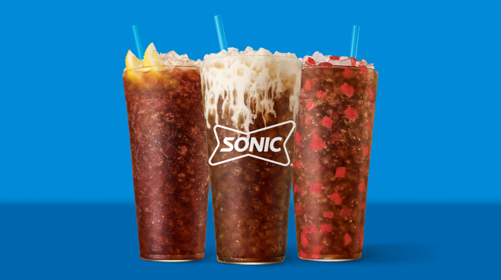 Three tall Sonic drinks filled with ice are shown: one with lemon slices, one with creamy topping, and one with red candy pieces, all in clear cups with blue straws against a blue background.