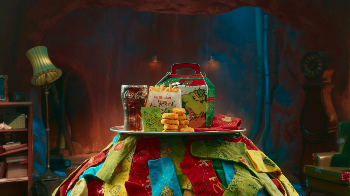 A tray with a Coca-Cola glass, chicken nuggets, and a festive takeout box sits atop a colorful patchwork blanket, inside a cozy, dimly lit room decorated with quirky furniture and warm lighting.