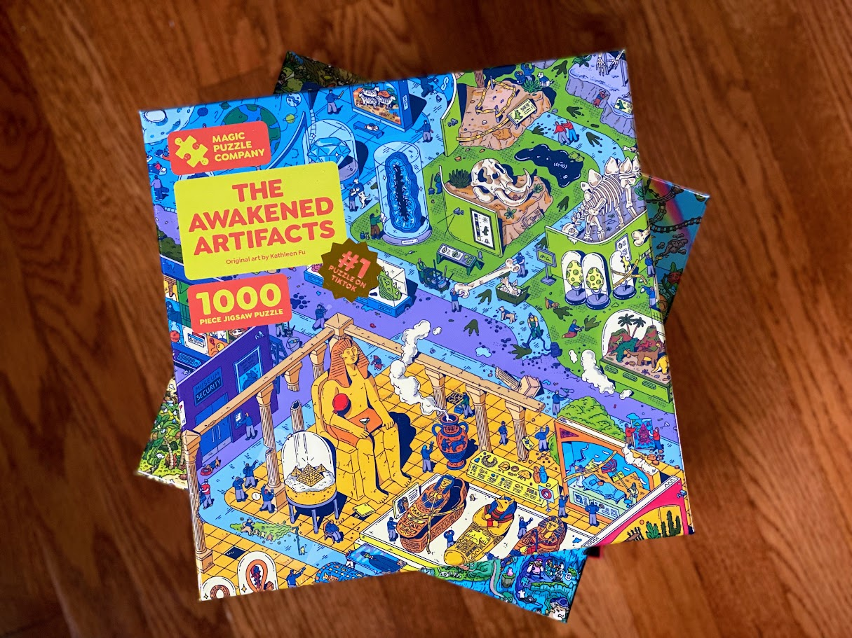 A colorful 1000-piece jigsaw puzzle box titled "The Awakened Artifacts" by Magic Puzzle Company sits on a wooden floor, featuring a busy illustrated scene with various rooms, artifacts, and characters.
