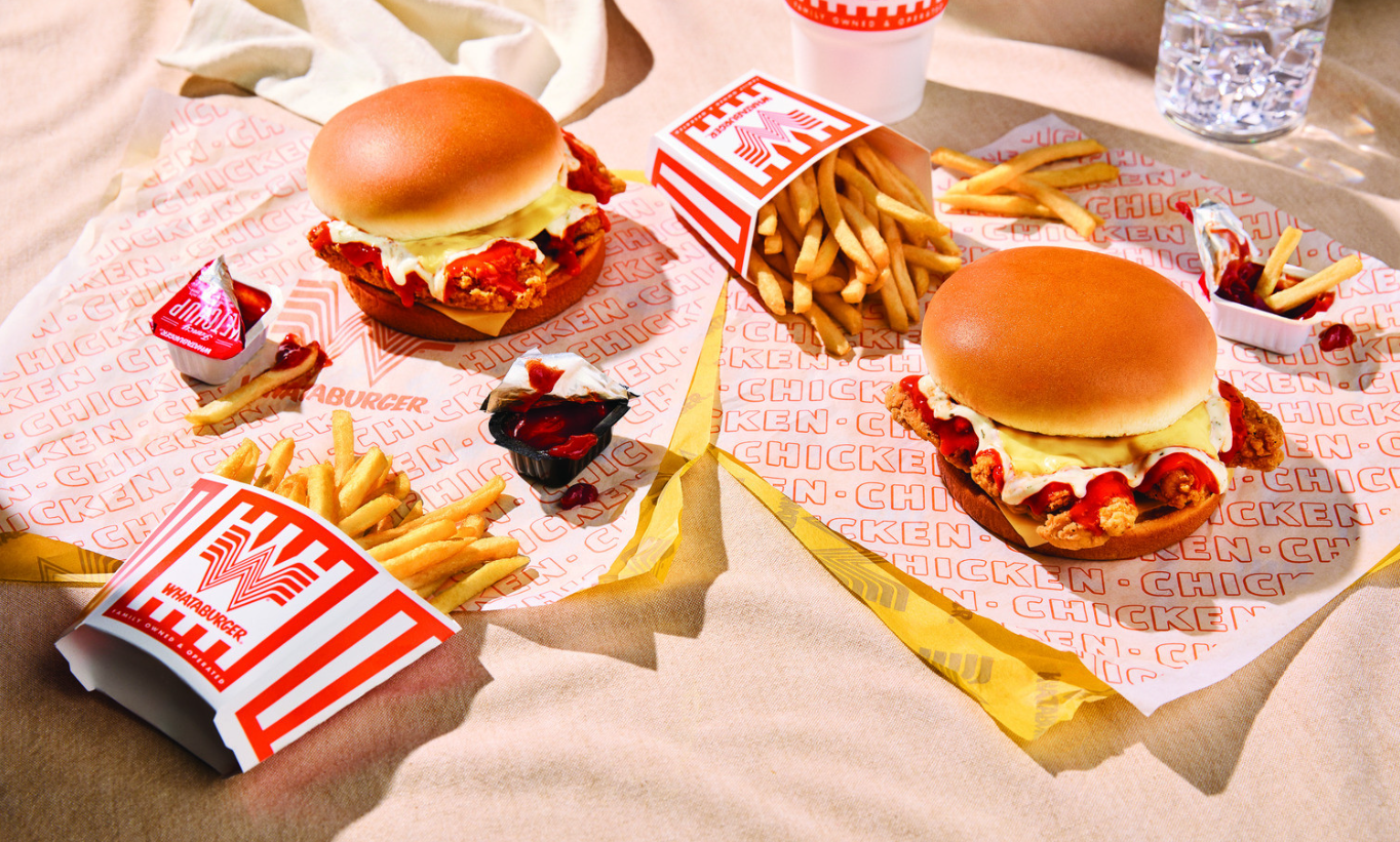 Two Whataburger chicken sandwiches with fries, ketchup packets, and a drink are placed on branded paper atop a sandy beige surface, presenting a casual picnic-style meal from the new fast-food menu.
