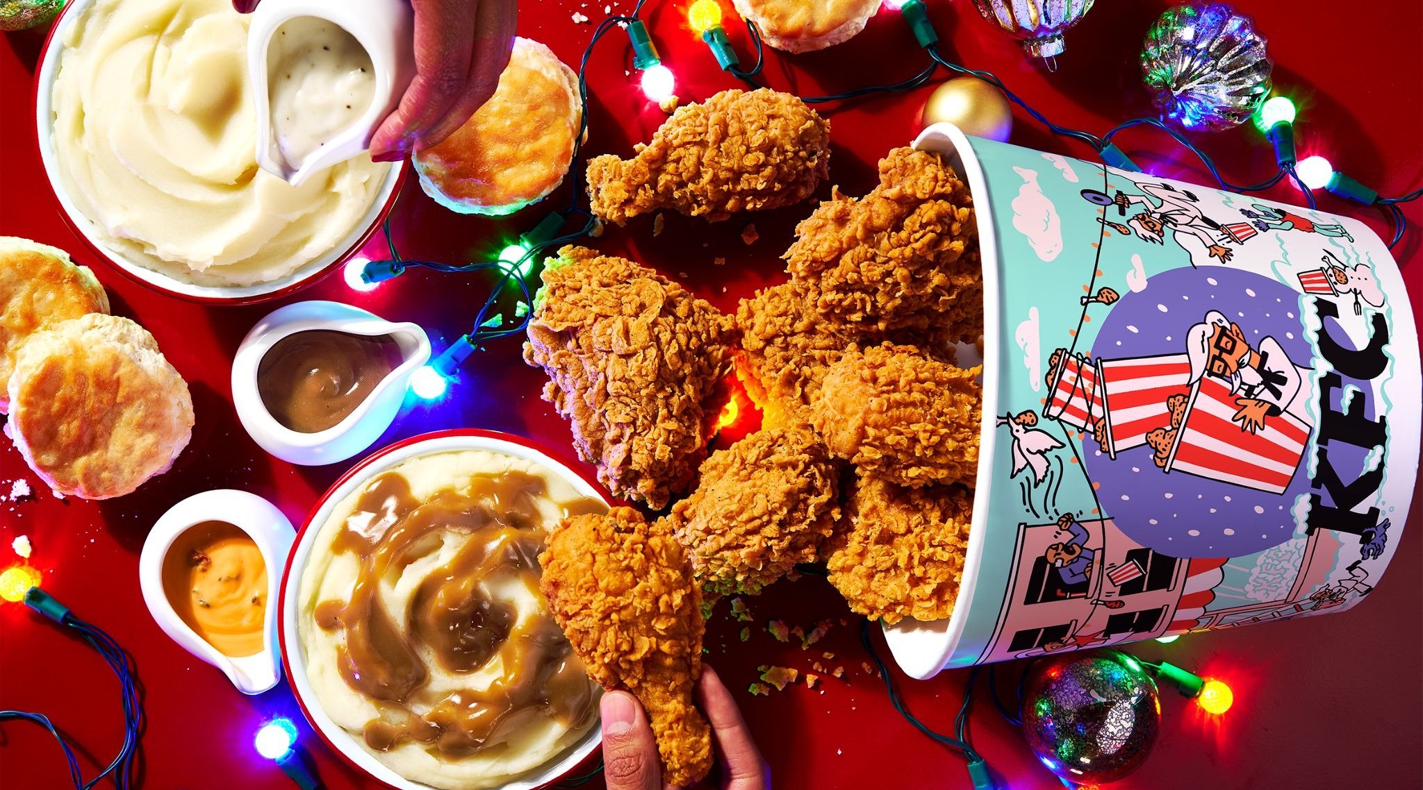 A festive table with a KFC bucket of fried chicken, mashed potatoes with gravy, biscuits, and dipping sauces, surrounded by colorful holiday lights on a red background.