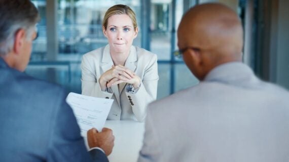 A woman in a light suit sits at a table with her hands clasped, looking nervous, while two people across from her, holding documents, appear to interview her in a modern office setting.
