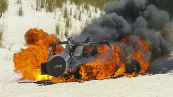 A Jeep is engulfed in intense flames and thick black smoke on a sandy area with sparse grasses in the background. The fire appears to be destroying the entire vehicle.