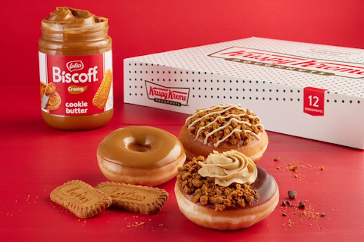 Three doughnuts with Biscoff cookie butter toppings sit in front of a jar of Lotus Biscoff spread and an open box of Krispy Kreme doughnuts on a red background, with Biscoff cookies scattered nearby.