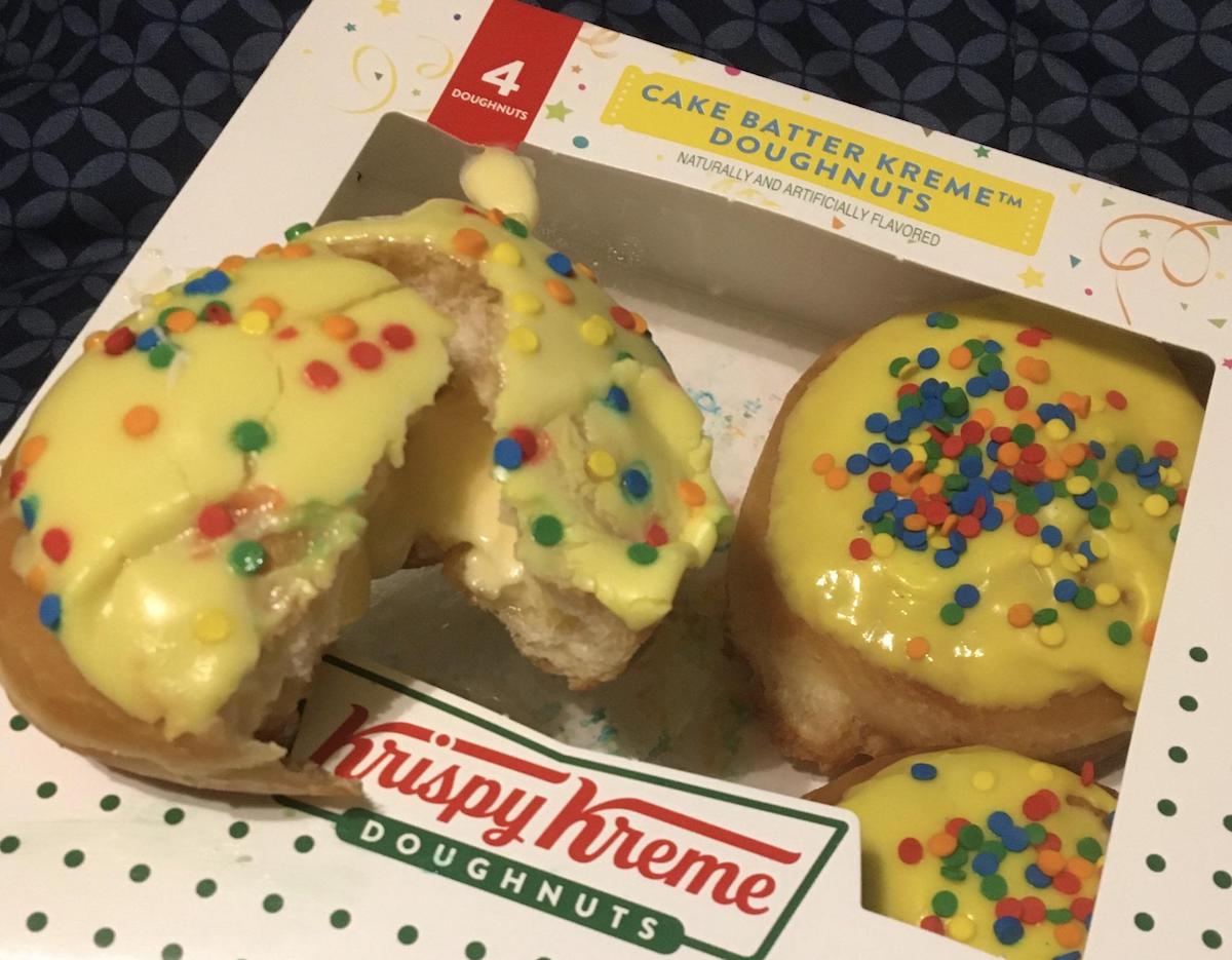 A box of four Krispy Kreme doughnuts, topped with yellow icing and colorful sprinkles. One doughnut is broken open, showing a creamy filling inside. The box label reads "Cake Batter Kreme Doughnuts.