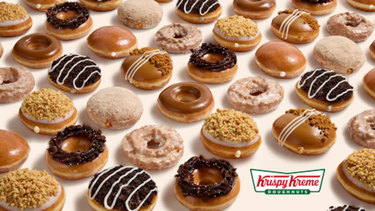 An assortment of Krispy Kreme doughnuts, including glazed, chocolate, nut-topped, and cream-filled varieties, are arranged in neat rows on a light background. The Krispy Kreme logo appears in the bottom right corner.