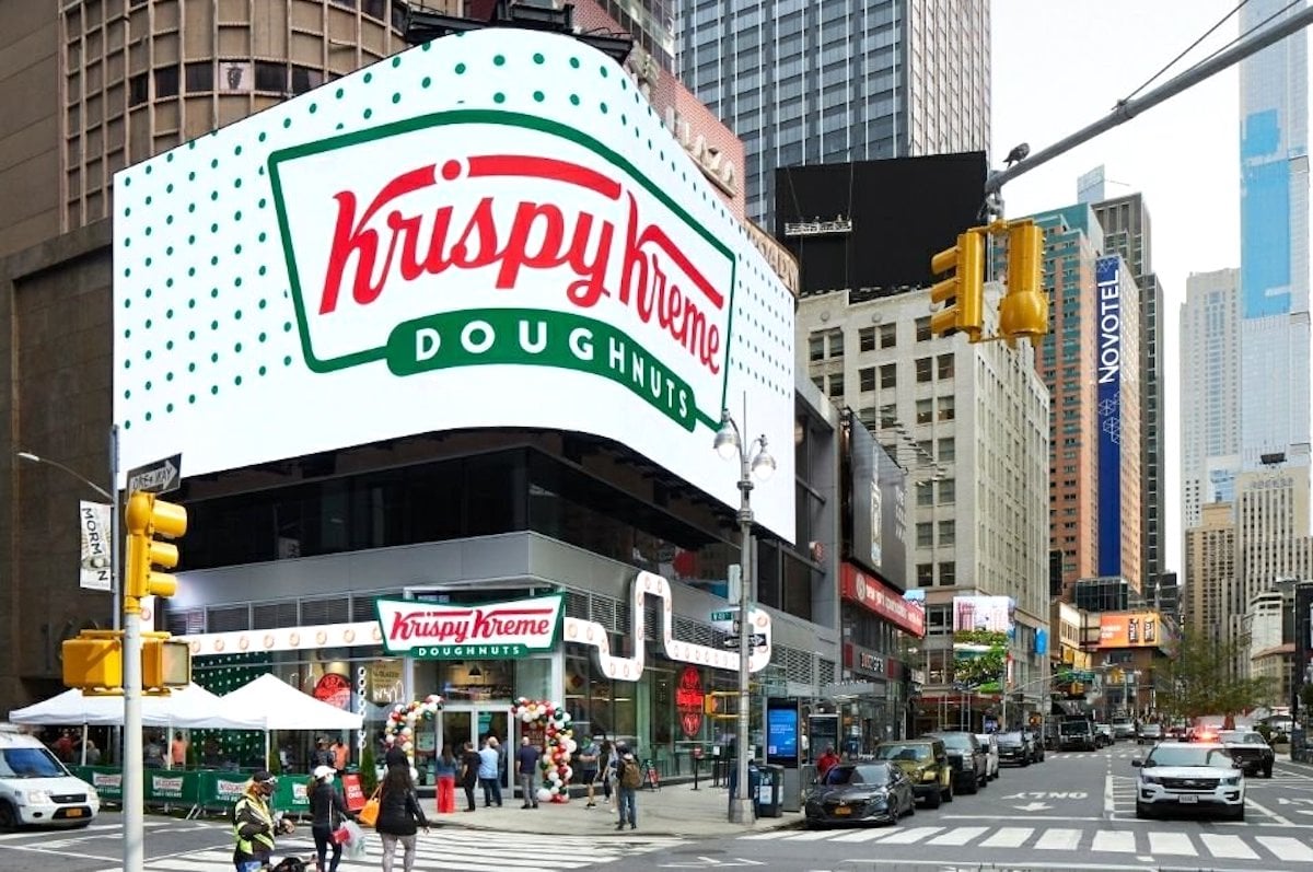 A Krispy Kreme Doughnuts store on a busy city street corner with a large digital sign above the entrance. People and cars are visible, and tall buildings surround the area.