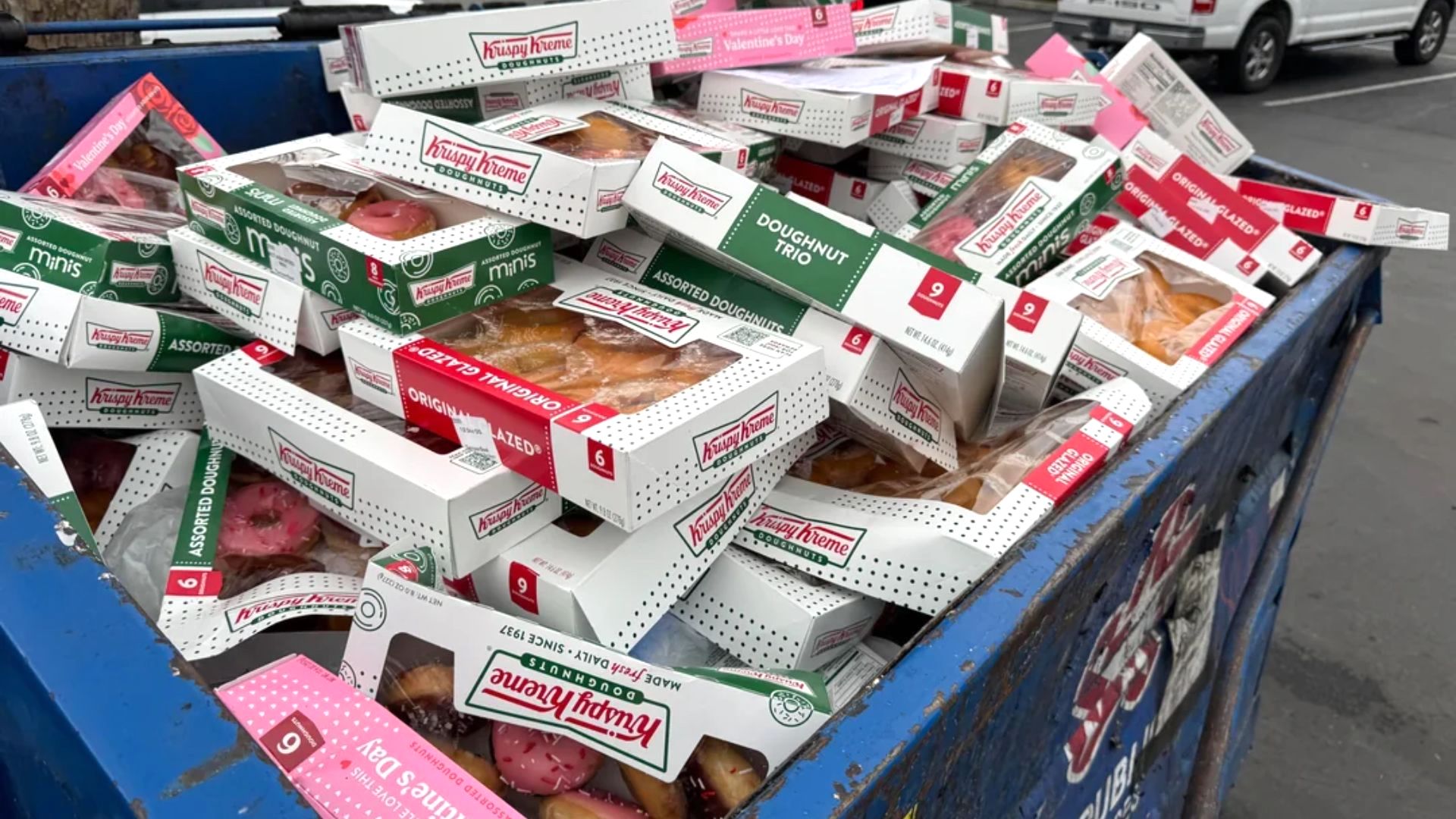A large pile of unopened Krispy Kreme donut boxes discarded in a blue dumpster outdoors, with various flavors visible through the clear windows on the boxes.