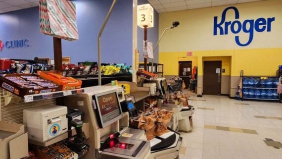Self-checkout area at a Kroger grocery store, with bags of groceries, candy displays, and a large blue Kroger logo on a yellow wall in the background.
