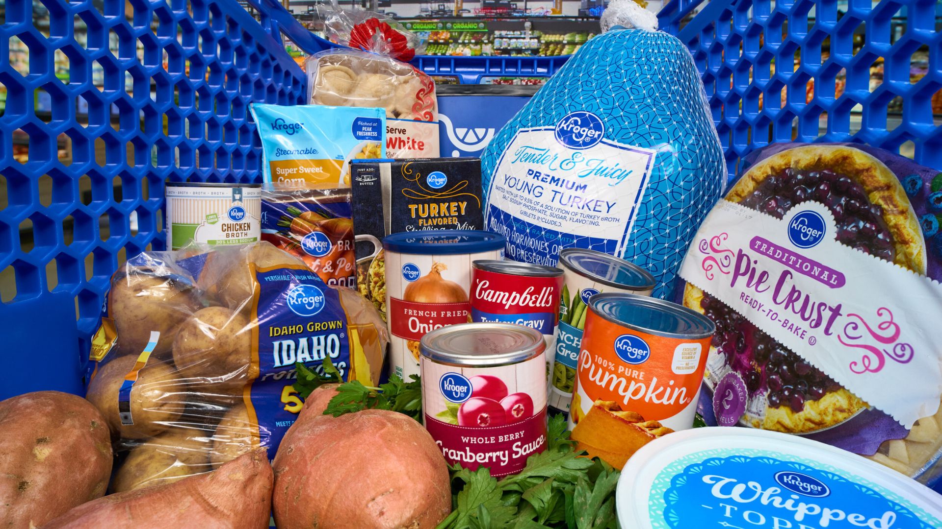 A grocery cart filled with Thanksgiving food items, including a frozen turkey, potatoes, sweet potatoes, canned pumpkin, cranberry sauce, pie crust, whipped topping, stuffing mix, bread, and vegetables.