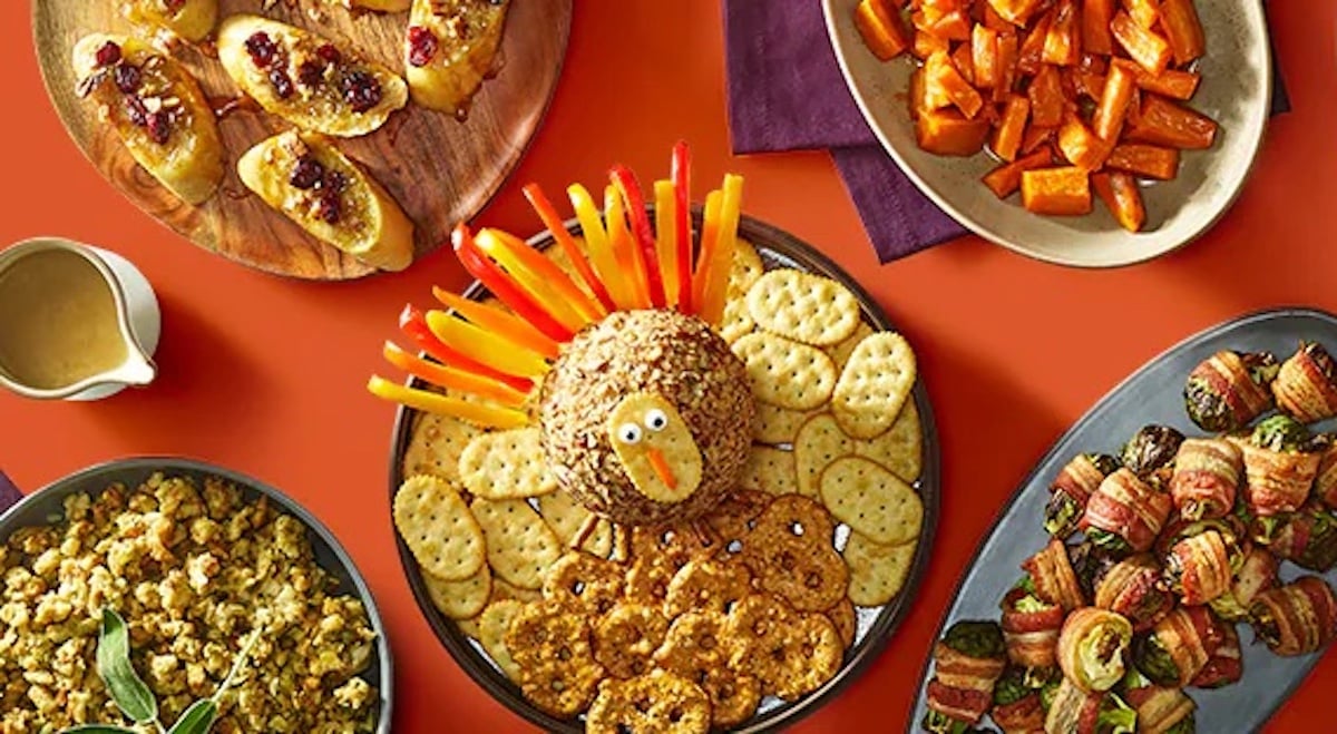 A festive spread of Thanksgiving appetizers, including a cheese ball shaped like a turkey with crackers, stuffed potato skins, roasted carrots, bacon-wrapped veggies, and a bowl of popcorn on an orange table.