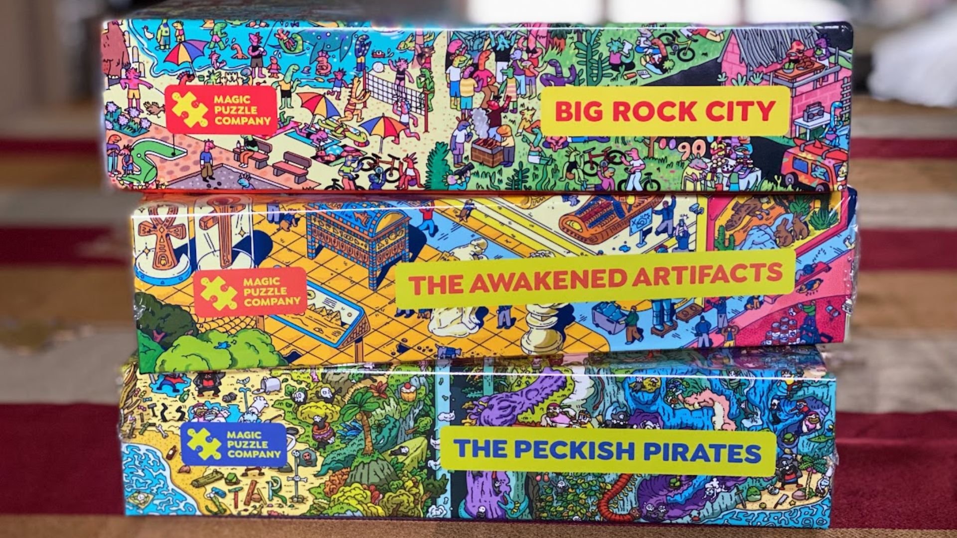 Three colorful puzzle boxes from the Magic Puzzle Company are stacked. The titles on the spines read: "BIG ROCK CITY," "THE AWAKENED ARTIFACTS," and "THE PECKISH PIRATES," in bright yellow blocks.