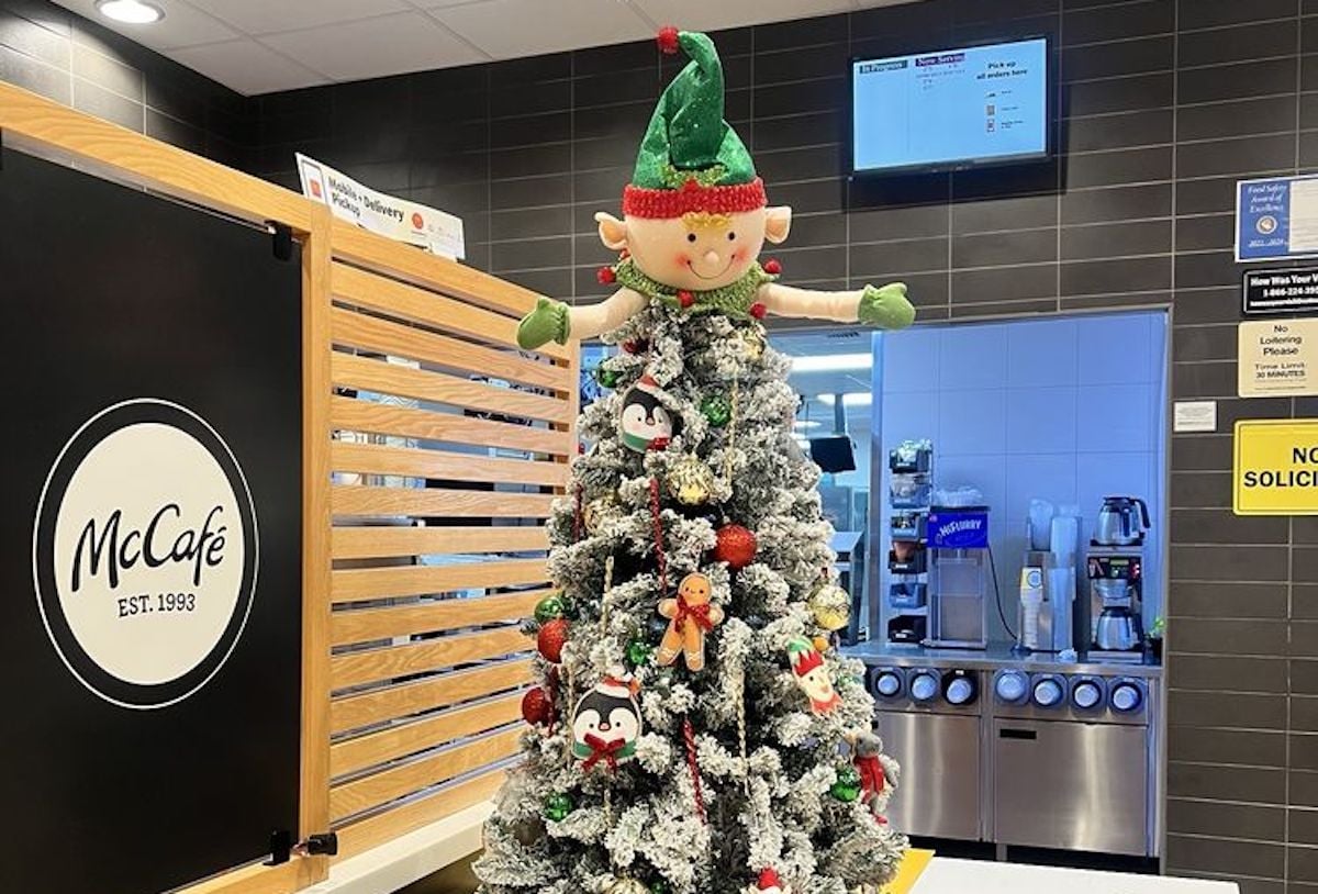 A Christmas tree decorated with snow-covered branches, ornaments, and topped with a large elf figure stands inside a McDonald's McCafé near a drinks station.