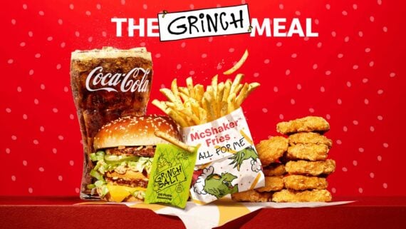 A meal with a large Coca-Cola, a cheeseburger, fries, chicken nuggets, and a green "Grinch Salt" packet on a red background, with "The Grinch Meal" text and Grinch illustrations on the packaging.