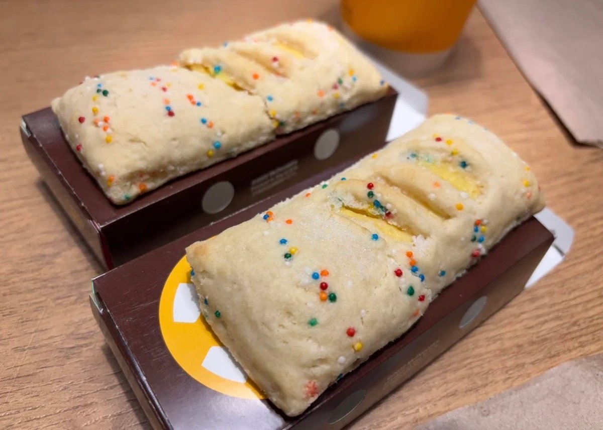 Two rectangular pastries with a light, flaky crust topped with colorful sprinkles are placed in brown cardboard trays on a wooden surface. A cup is partially visible in the background.
