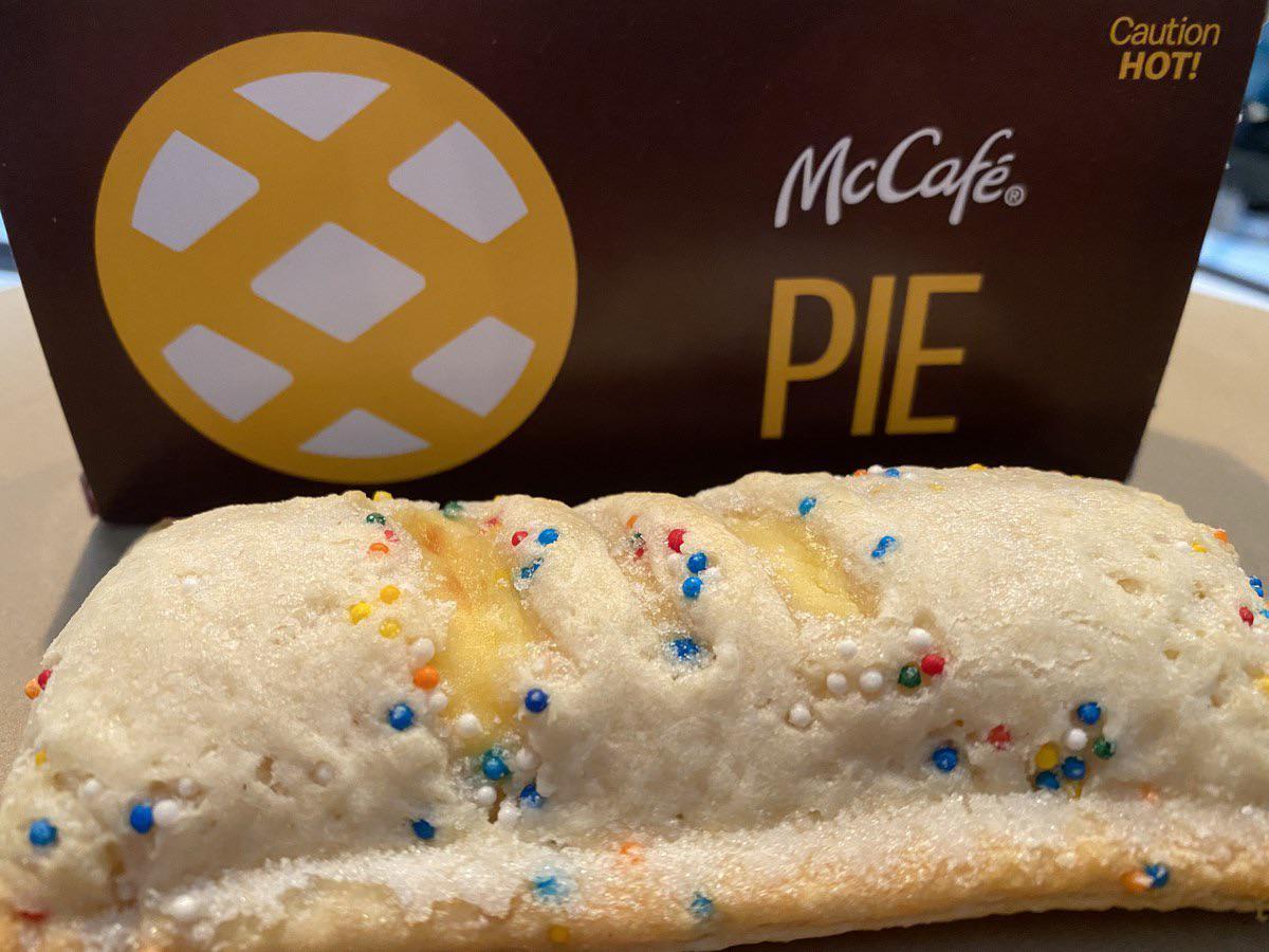 A McDonald's McCafé pie with a golden crust and colorful sprinkles sits in front of its brown and yellow pie box, which has a lattice pie graphic and the word "PIE" printed on it.