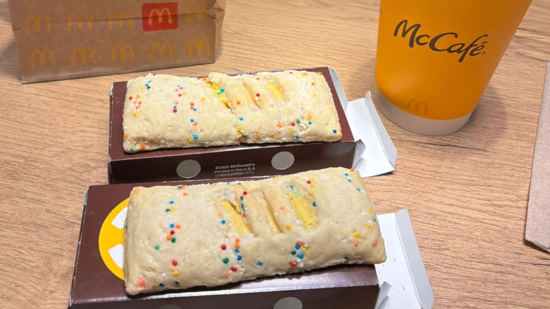 Two McDonald's dessert pies with sprinkle topping sit in open boxes on a wooden table next to a McCafé cup and a paper McDonald's bag.