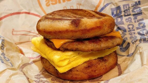 A breakfast sandwich with sausage, scrambled egg, and melted cheese between two griddled pancake-style buns, wrapped in branded paper.