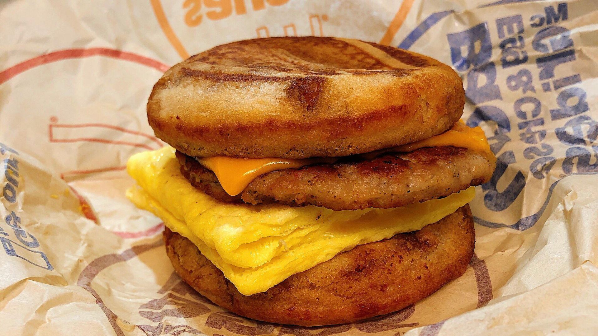 A breakfast sandwich with sausage, scrambled egg, and melted cheese between two griddled pancake-style buns, wrapped in branded paper.