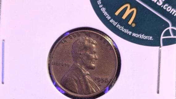 A 1956 D Lincoln penny displayed in a white holder, with part of a green and yellow McDonald's promotional sticker visible in the upper right corner.