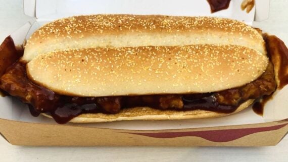 A sandwich with a saucy boneless rib patty in a long sesame seed bun, served in an open cardboard container. BBQ sauce drips from the meat, slightly spilling over the sides of the bread.