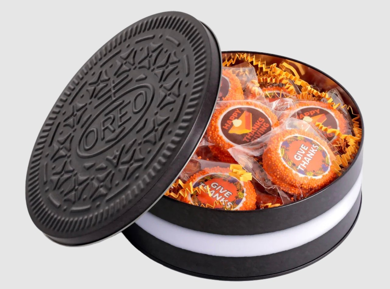 A round container shaped like an Oreo cookie holds festive cookies decorated with orange sprinkles and messages like “HAPPY THANKSGIVING” and “GIVE THANKS.” The cookies sit in yellow shredded paper.