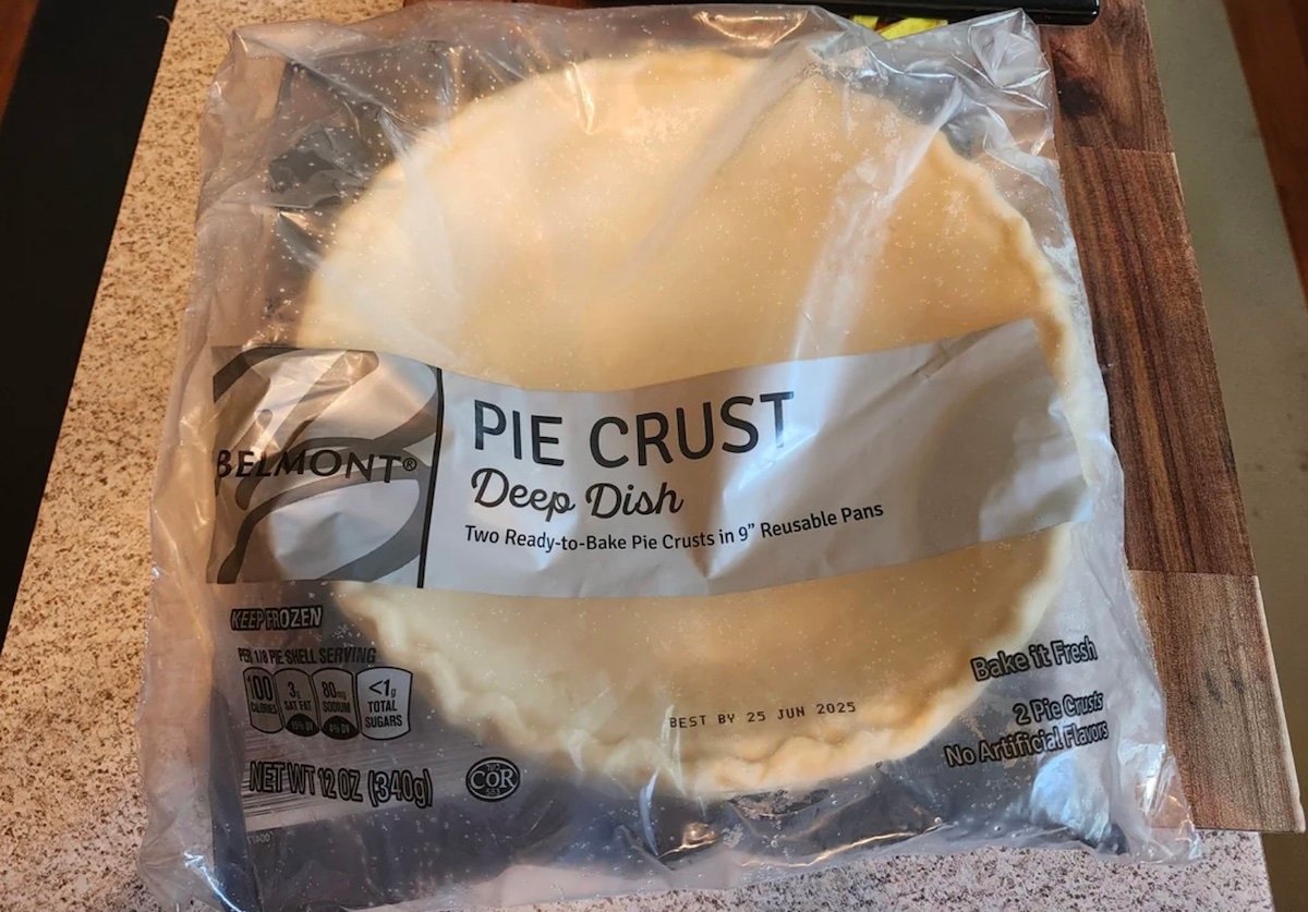 A package of Belmont brand deep dish pie crusts sits on a counter. The label states "Pie Crust Deep Dish," "Two Ready-to-Bake Pie Crusts in 9'' Reusable Pans," and the package is sealed and partially transparent.