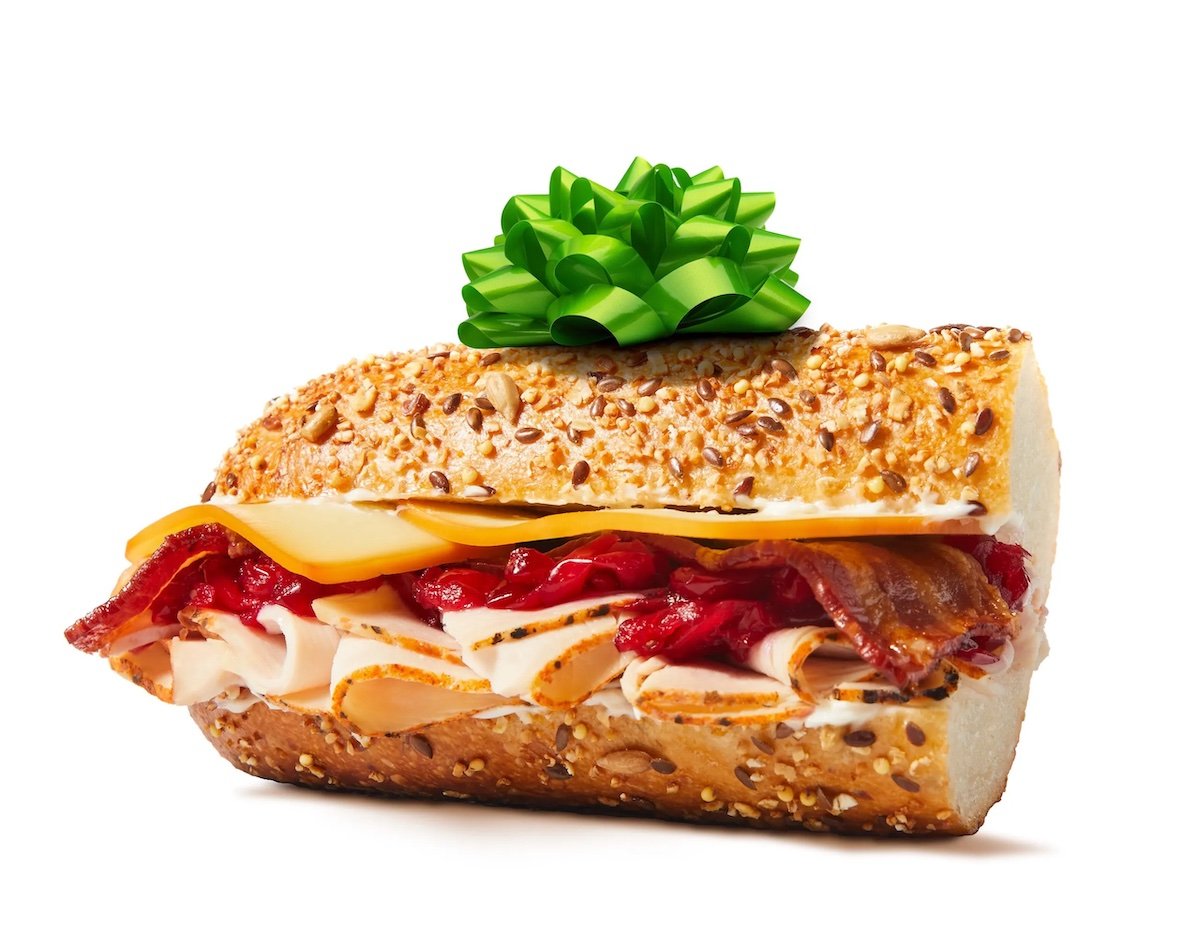 A sub sandwich with turkey, bacon, cheese, and cranberry sauce on seeded bread is topped with a green gift bow, photographed on a white background.