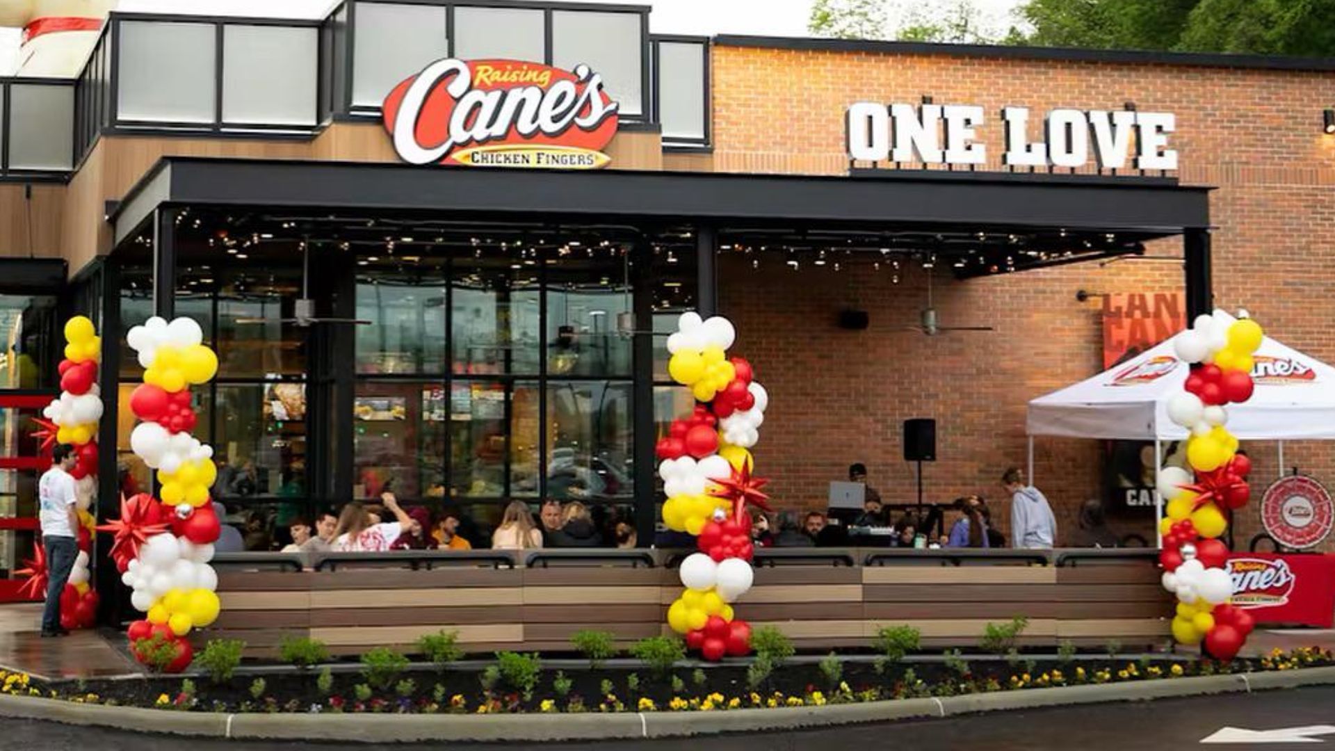 A Raising Cane’s restaurant with a patio decorated with red, white, and yellow balloons. The sign reads “ONE LOVE” above the entrance, and people are seated outside under string lights.