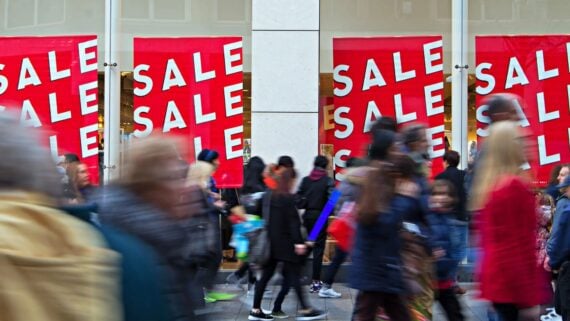 Large red "SALE" signs in store windows as a crowd of people walk past on a busy street, some blurred by motion, suggesting a bustling shopping scene during a retail sale event.