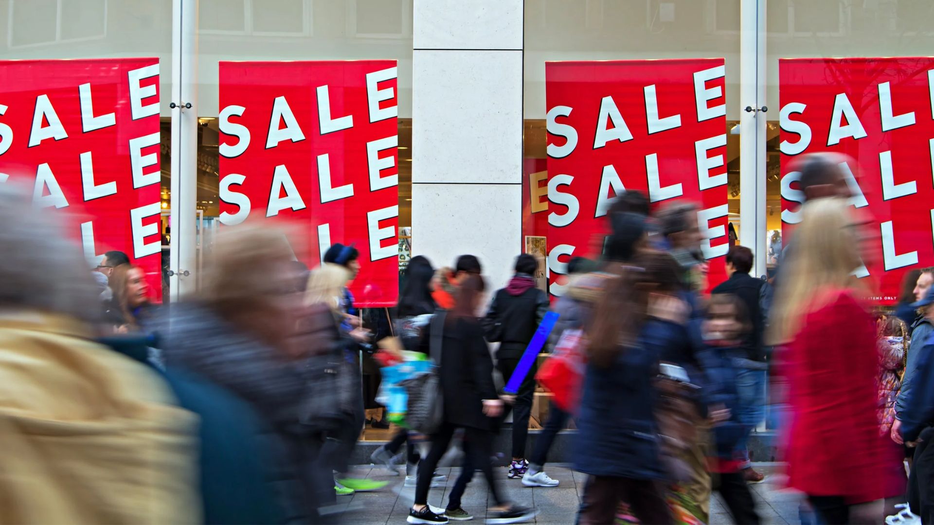Large red "SALE" signs in store windows as a crowd of people walk past on a busy street, some blurred by motion, suggesting a bustling shopping scene during a retail sale event.