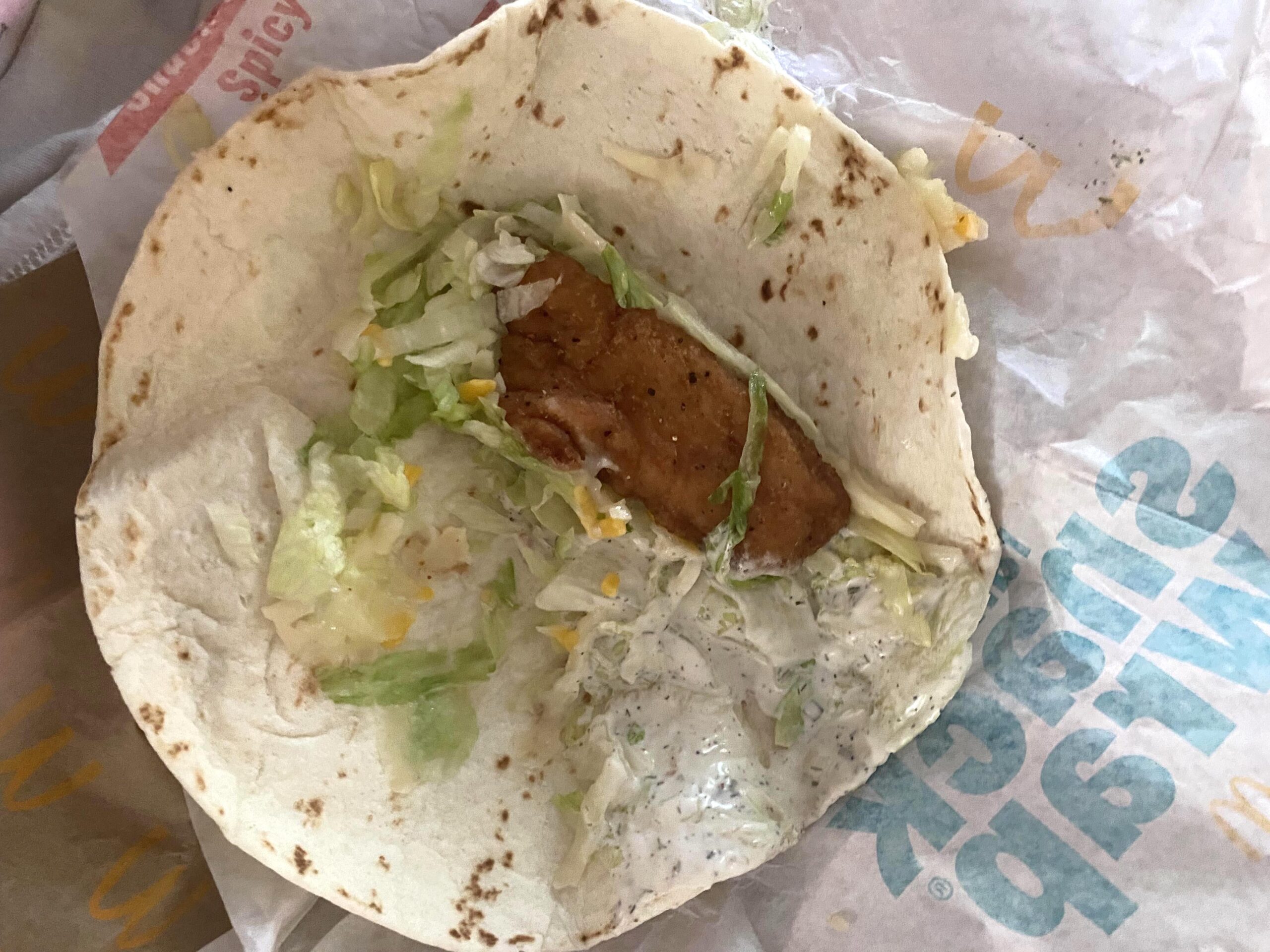 A soft flour tortilla with a piece of breaded chicken, shredded lettuce, shredded cheese, and ranch sauce, partially open on branded wrapper paper.
