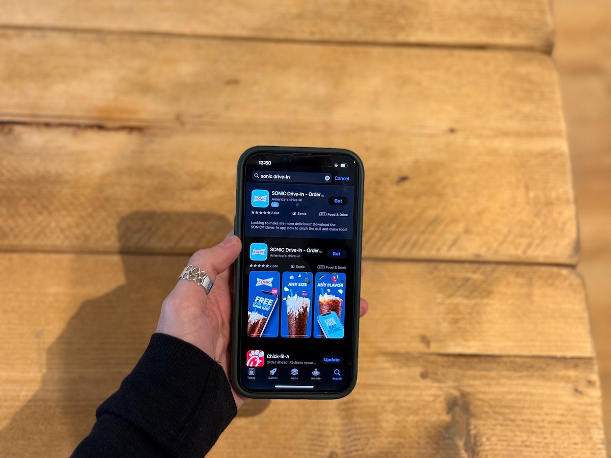 A hand holding a smartphone displaying the SONIC Drive-In app page in the App Store, with a wooden table background.
