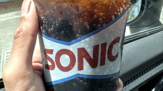 A hand holds a clear plastic cup filled with a dark, carbonated beverage. The cup has the SONIC logo on it, and condensation is visible on the outside of the cup. The background shows part of a car window.