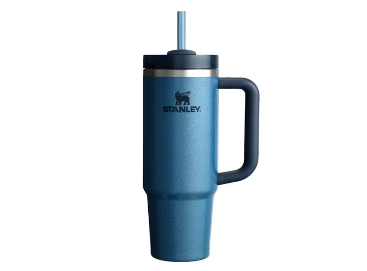 A blue Stanley travel tumbler with a handle, lid, and straw is shown upright against a white background. The cup features the Stanley logo on the front.