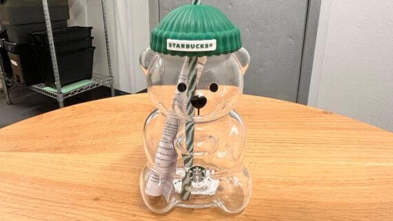 A clear plastic bear-shaped container with a green Starbucks beanie lid sits on a wooden table. Inside, there is a straw and some paper. The container has a Starbucks logo on its side.