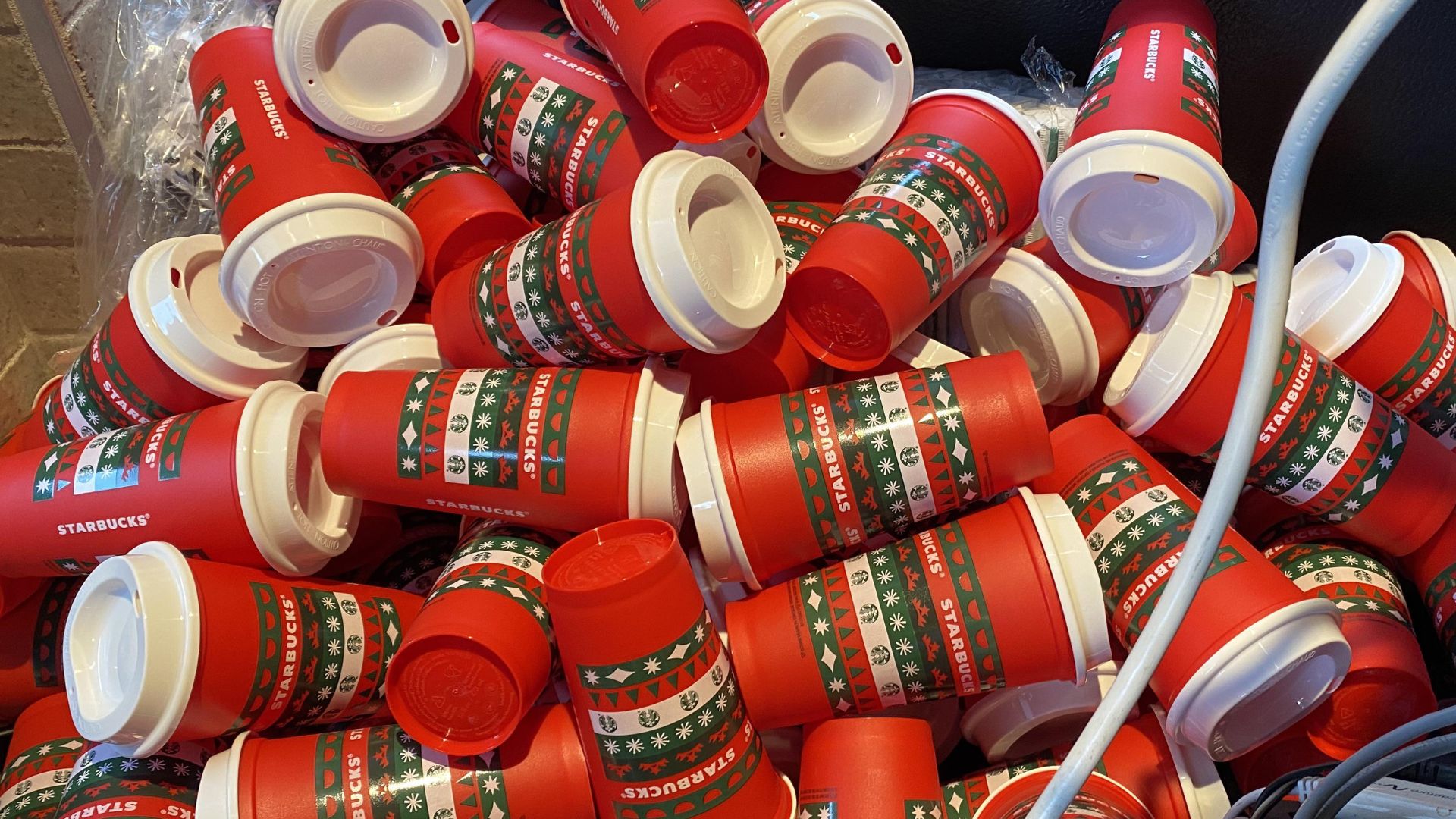 A large pile of red Starbucks holiday-themed cups with white lids, decorated with green and white festive patterns, stacked haphazardly together.