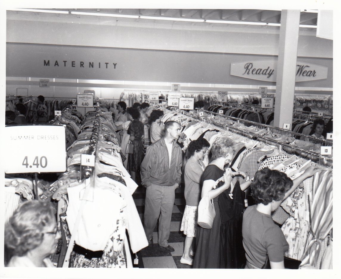 Black-and-white photo of people shopping for clothes in a crowded store. Signs mark sections like "Maternity" and "Ready to Wear." A sign displays "Summer Dresses 4.40." Women browse racks of clothing.