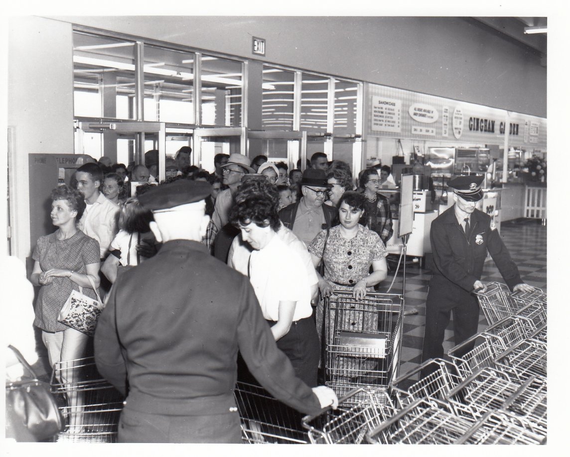 Black and white photo of a crowd entering a supermarket, with men in uniforms managing grocery carts. People, mostly women, hold baskets or push carts as they move through the store’s entrance area. Signs hang on the walls.