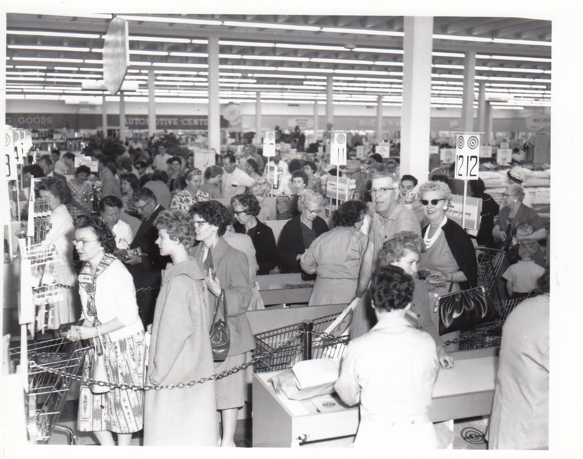 Black-and-white photo of a crowded supermarket with many women waiting in line at checkout counters. Shopping carts are filled with groceries, and the store has bright fluorescent lights overhead.