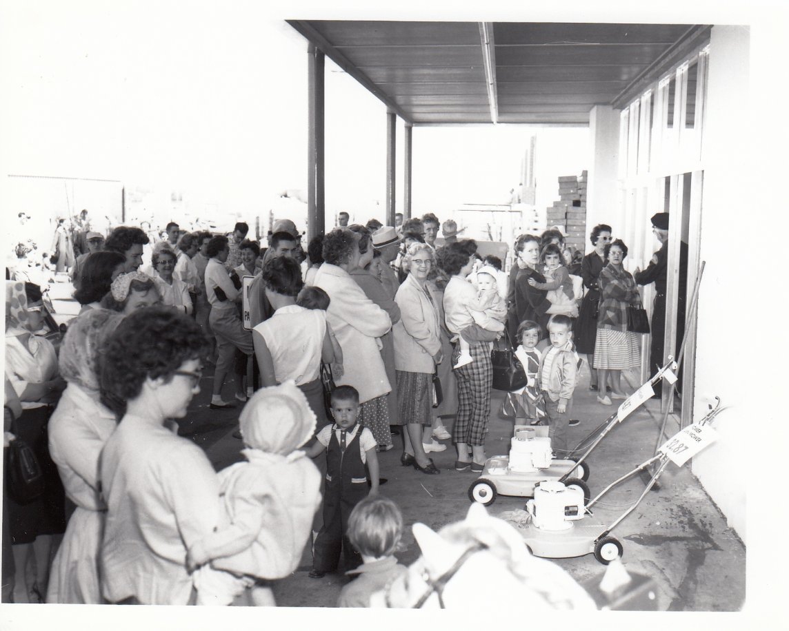 A large group of women and children gather outside a building, some holding infants. A couple of lawn mowers are visible in the foreground. The scene appears busy and social, with people standing and talking.