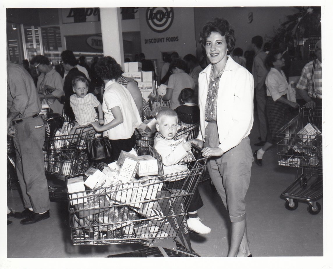 A woman smiles while pushing a shopping cart filled with groceries and a young child at a busy supermarket. Other shoppers with carts and children are visible in the background. The image appears to be from the mid-20th century.