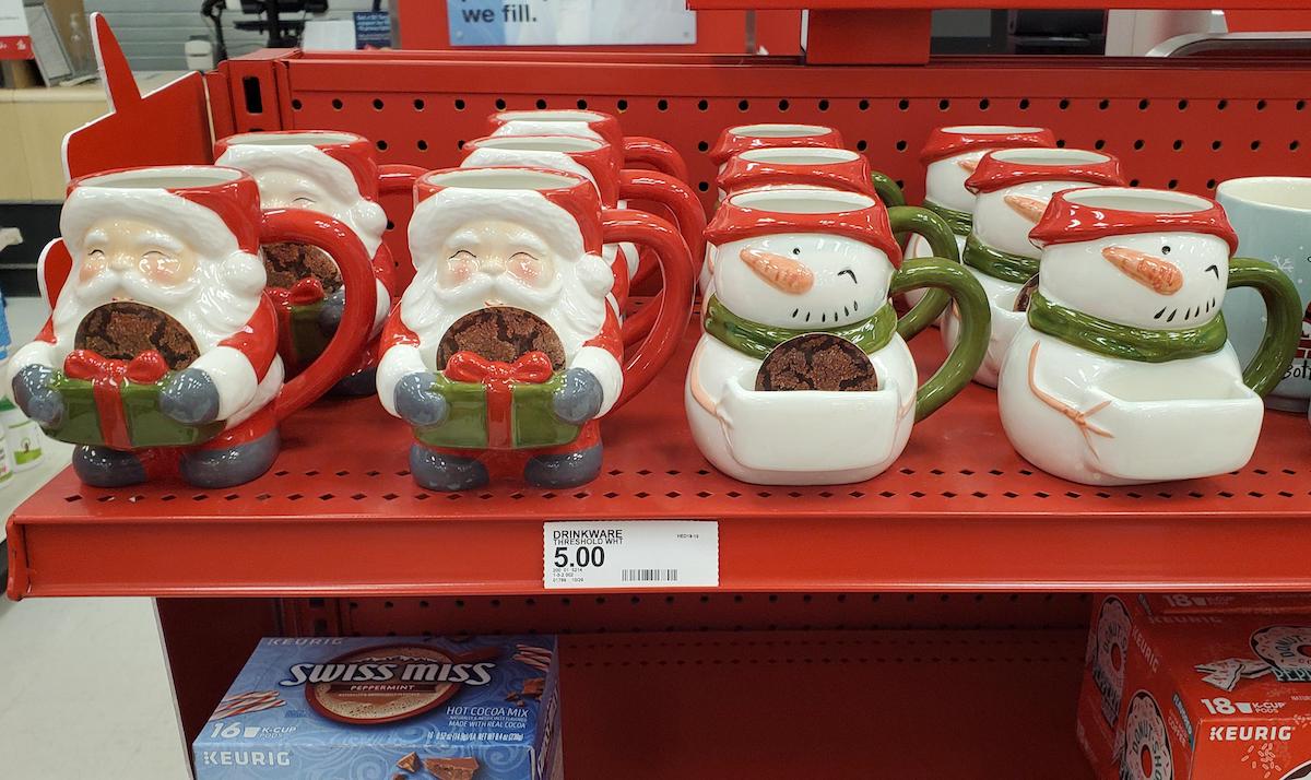 Ceramic Santa Claus and snowman mugs are displayed on a red store shelf. Each mug holds a cookie where the mouth would be. A price tag shows they are $5. Boxes of Swiss Miss hot chocolate are on a lower shelf.