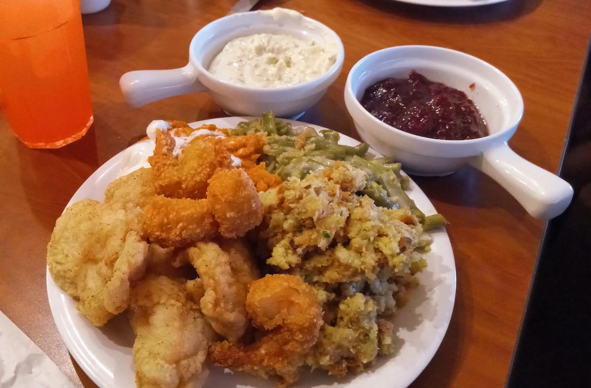 A plate of fried shrimp, fried fish, stuffing, and green beans, served with bowls of tartar sauce and cranberry sauce on a wooden table. An orange drink is partially visible on the side.