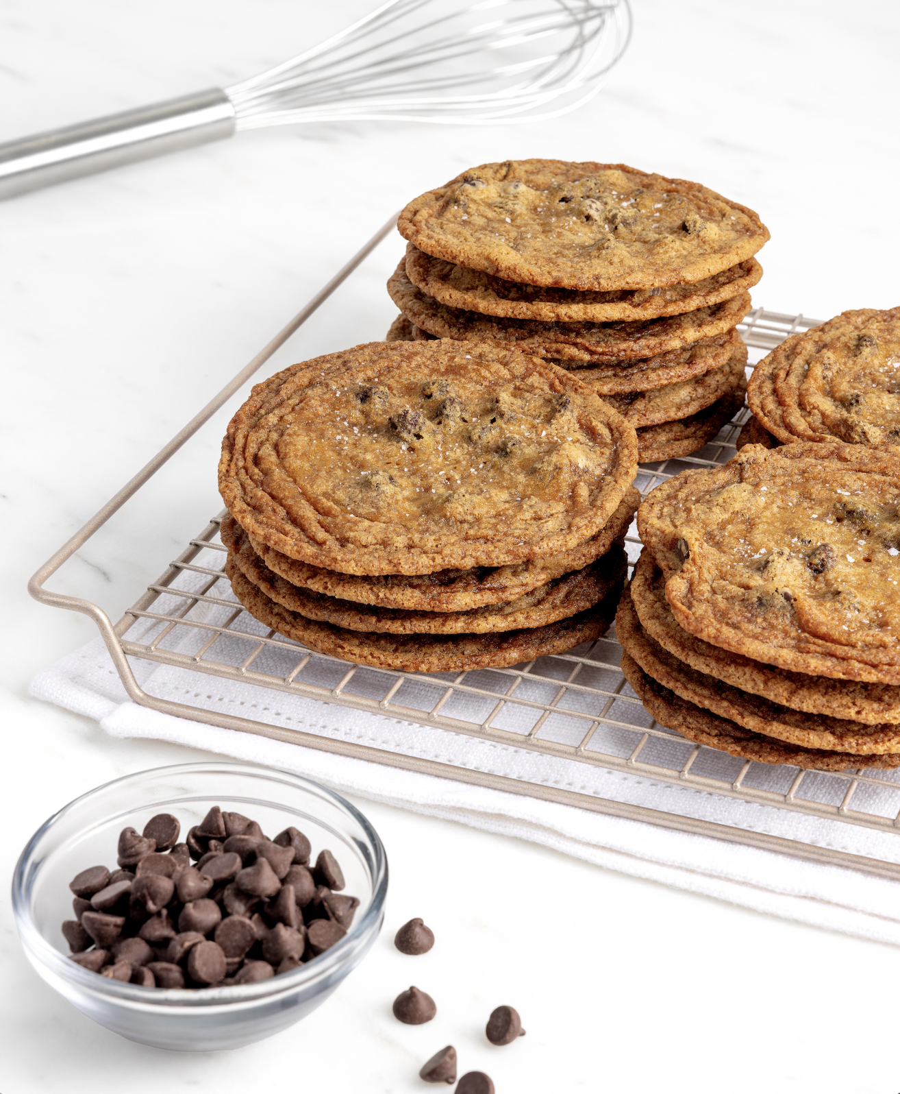 Three stacks of chocolate chip cookies rest on a cooling rack, with a glass bowl of chocolate chips and a whisk nearby on a white surface.