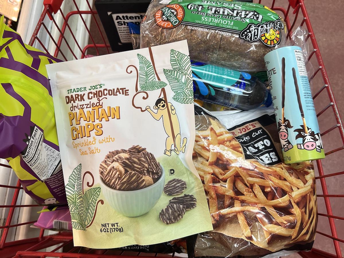 A red shopping basket filled with groceries, including Trader Joe’s Dark Chocolate Drizzled Plantain Chips, a loaf of bread, a bag of chips, packaged fries, deodorant, and other assorted food items.