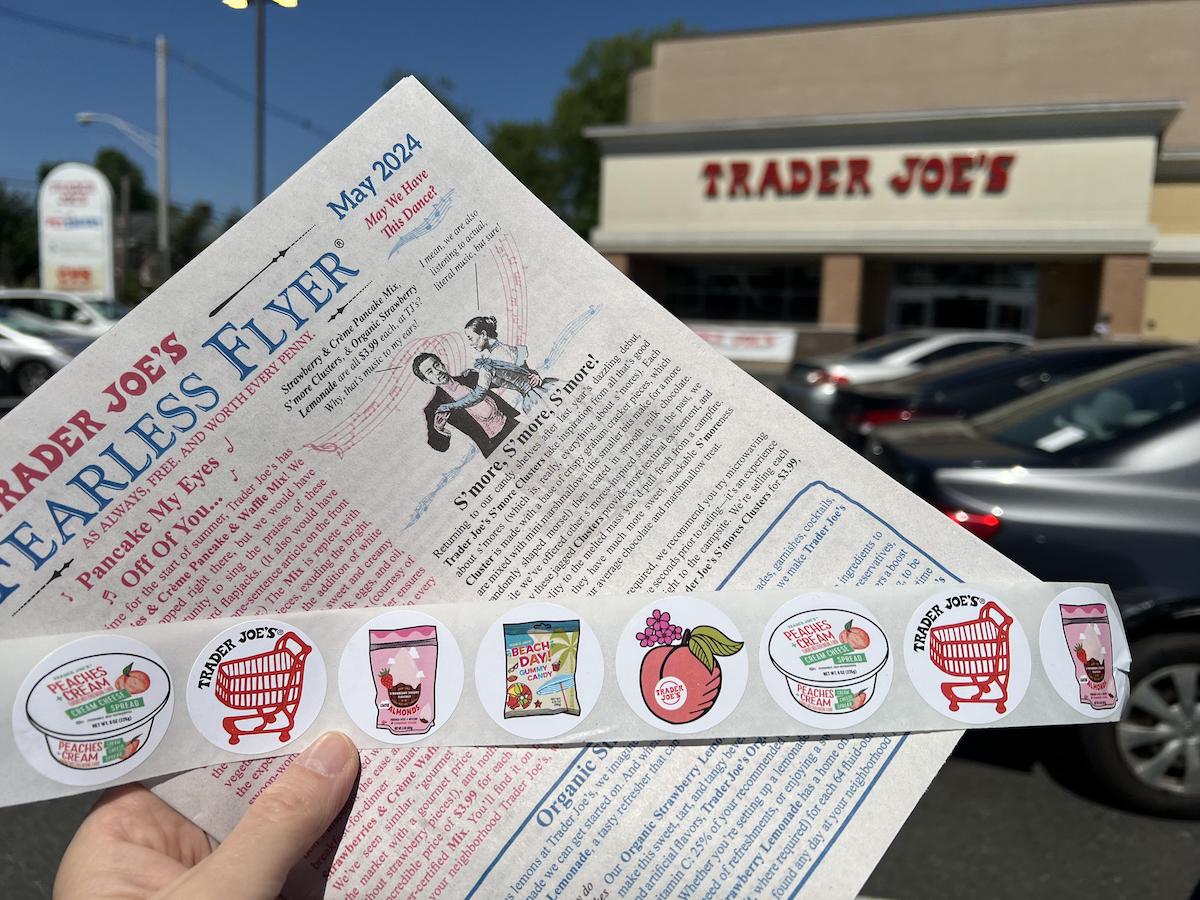 A hand holds a Trader Joe’s Fearless Flyer newsletter and a strip of Trader Joe’s stickers outside a Trader Joe’s store, with parked cars and store signage visible in the background.
