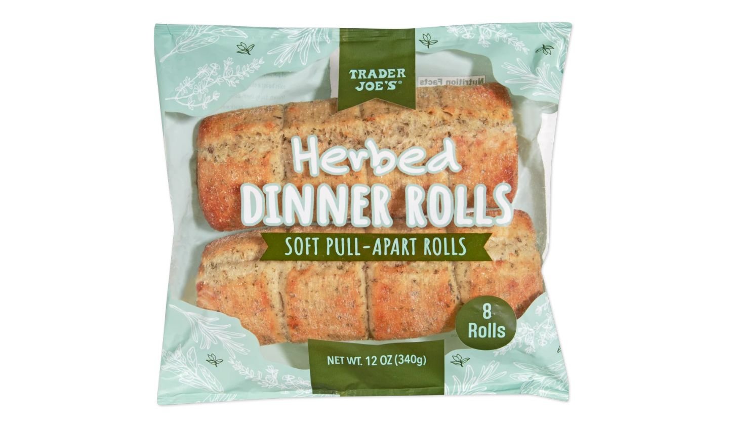 A package of Trader Joe’s Herbed Dinner Rolls, labeled “Soft Pull-Apart Rolls,” contains 8 rolls and weighs 12 oz (340g). The packaging is green with white floral designs and a clear window displaying the rolls.