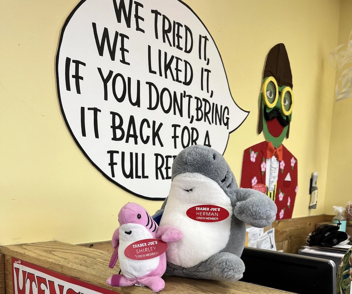 Two plush toys, a gray shark and a pink narwhal, with name tags labeled “Trader Joe’s Herman Chominsky” and “Trader Joe’s Shirley Chompstein,” sit in front of a speech bubble sign about returns. A colorful cutout is in the background.