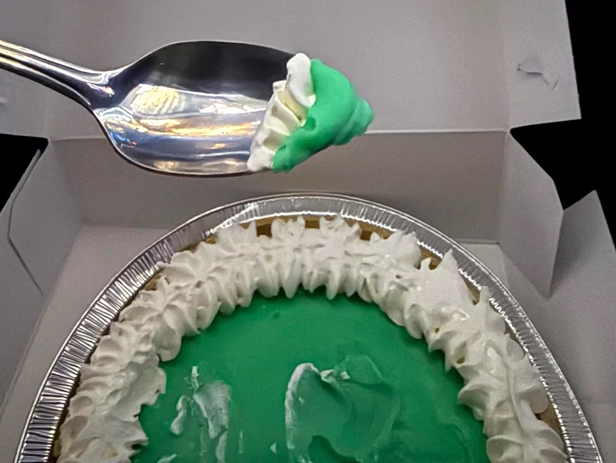 A spoon holds a bite of green pie with whipped cream above a larger pie in a foil tin. The pie has a bright green filling and a whipped cream border, and sits in an open white box.
